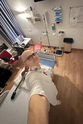 Persona con gafas y bata médica en una habitación con cama hospitalaria, guitarra, computadora portátil, silla y soporte de suero