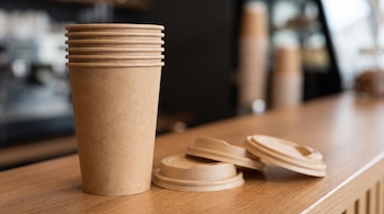 Una pila de vasos de papel marrón sin texto y tres tapas sobre un mostrador de madera en una cafetería con fondo desenfocado.