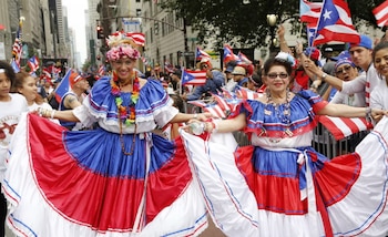 Dos mujeres sonrientes con trajes folclóricos puertorriqueños, azul, rojo y blanco, de faldas amplias. Llevan tocados florales y collares. Desfile con banderas y gente