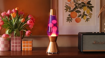 Primer plano de una lámpara de lava dorada y morada encendida junto a tulipanes rosados y naranjas en jarrones, un altavoz Marshall negro y un cuadro frutal enmarcado.