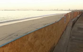 Playa de arena con el océano, una barrera de madera y un muro de concreto, una ciudad distante y un cielo brumoso