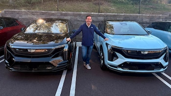 Un hombre de pie en un estacionamiento entre dos automóviles de la marca Cadillac, uno negro y otro celeste