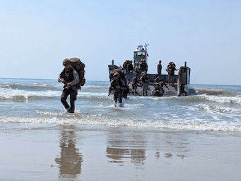 Militares con uniformes de camuflaje y grandes mochilas corren a través de las olas poco profundas desde un buque de desembarco hacia la playa bajo un cielo azul claro