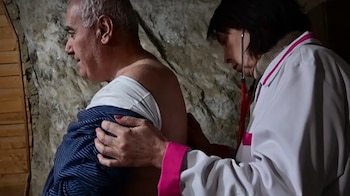 Una mujer vestida con bata blanca y estetoscopio examina la espalda desnuda de un hombre mayor sentado. Se ve una pared rocosa