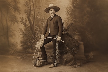 Fotografía sepia de un hombre con sombrero, chaqueta, pantalón decorado y bigote que sostiene una soga y apoya un pie sobre una piedra.