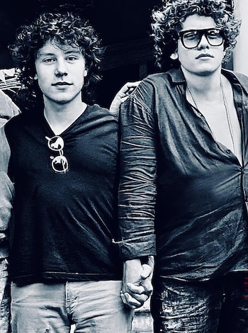 Dos hombres jóvenes de cabello rizado posan tomados de la mano. Uno lleva gafas colgando de su camiseta y el otro gafas oscuras puestas