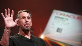 Un hombre de mediana edad con barba, Larry Ellison, saluda con la mano izquierda levantada. Viste una camisa oscura. Al fondo, un letrero borroso con el logo de Oracle
