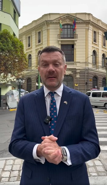 Richard Porter, un hombre caucásico de mediana edad con barba, viste un traje azul a rayas y corbata. Está de pie en una calle urbana con un cruce peatonal y edificios antiguos de fondo