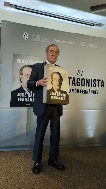 José Ramón Fernández, un hombre mayor con traje oscuro y gafas, de pie frente a un banner, sostiene su libro "El Protagonista" con su imagen en la portada