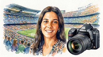 Mujer sonriendo con cabello oscuro, blusa estampada, un estadio de fútbol con gradas llenas de público y una cámara réflex Nikon D780.