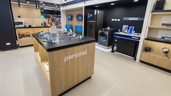 Interior de tienda de electrónica. A la derecha, refrigerador y lavadora Samsung negros. Al centro, mostrador "Personal" con smartphones. Al fondo, televisores