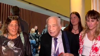 El médico Alberto Cormillot viste traje y corbata mientras permanece acompañado por varias personas en un vestíbulo