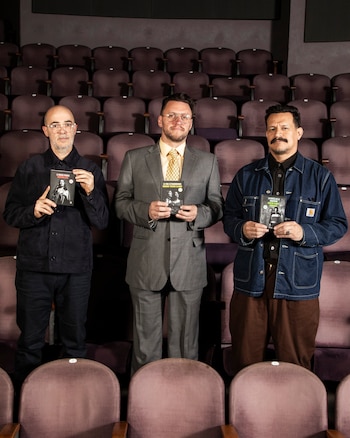 Tres hombres, uno calvo con gafas, uno con traje gris y gafas, y otro con chaqueta vaquera, posan de pie en un auditorio vacío sosteniendo programas