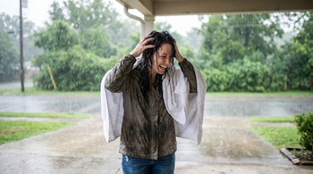 Mujer sonriente con cabello mojado se seca con una toalla blanca bajo un porche, con lluvia intensa visible al fondo y árboles verdes.