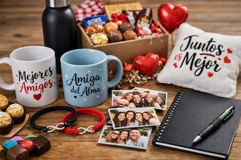 Mesa de madera con dos tazas, pulseras, fotos impresas, cuaderno, bolígrafo, botella térmica, caja de chocolates, almohadón y decoración de corazones.
