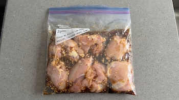Bolsa de plástico con cierre sobre una superficie gris, llena de trozos de pollo crudo en salsa con ajo picado y una etiqueta escrita.
