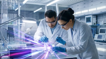 Dos científicos con batas, guantes y gafas manipulan un cristal facetado con pinzas sobre una mesa de laboratorio. El cristal dispersa haces de luz de colores.