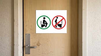 Cartel informativo en puerta de madera con figura humana sentada en inodoro dentro de círculo verde y figura de pie frente a inodoro dentro de círculo rojo tachado.