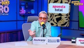 Un hombre con lentes de color naranja habla frente a un micrófono sentado frente a un escritorio con letreros