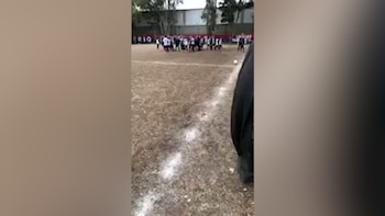 Terreno de juego con césped seco y línea blanca. Grupo de personas con uniformes deportivos en el centro. Una pelota de fútbol. Parte de ropa oscura en primer plano derecho