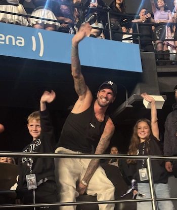 Un hombre con gorra negra y dos niños levantan la mano y sonríen desde la barra de un palco en un recinto con espectadores
