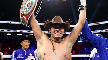 Un boxeador sin camiseta lleva un sombrero de vaquero y sostiene un cinturón de campeonato con los brazos en alto.