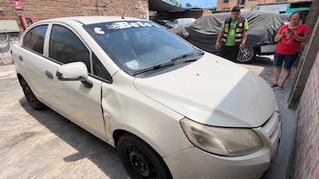 Un automóvil blanco con daños en la carrocería en un patio junto a un hombre con chaleco policial y una mujer con teléfono móvil