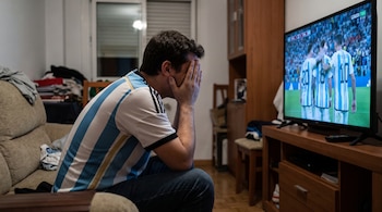Hombre sentado en un sofá con camiseta de Argentina cubre su cara, mirando un televisor que muestra a futbolistas de la selección argentina.