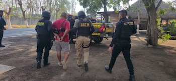 Tres agentes de policía escoltan a un hombre esposado que viste una camisa roja y pantalones cortos grises, junto a un vehículo policial y árboles