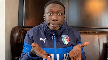 Un hombre joven de piel oscura con una chaqueta deportiva azul de Italia y logo de Puma, mirando a la cámara con una expresión de encogimiento de hombros y manos abiertas.