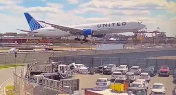 Un avión de pasajeros blanco y azul de United Airlines desciende para aterrizar sobre una carretera con tráfico y un estacionamiento con vehículos