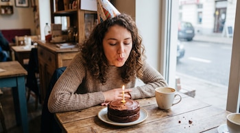 Mujer joven con cabello rizado y gorro de cumpleaños sopla una vela en un pastel de chocolate sobre una mesa de madera, con una taza de café al lado y una ventana al fondo.