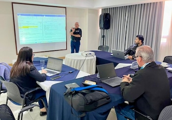 Un hombre presenta frente a una pantalla con software de Microsoft 365. Tres asistentes usan laptops en mesas con manteles azules en una sala de reuniones