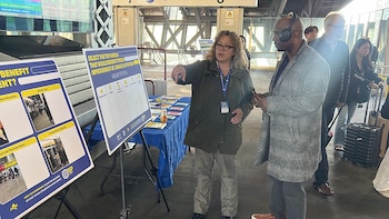 Una mujer con gafas y chaqueta señala un panel informativo azul sobre mejoras en la estación Jamaica a un hombre con auriculares, en un vestíbulo con pasajeros