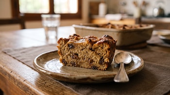 Una porción de budín de pan con pasas de uva sobre un plato de cerámica con una cuchara en una mesa de madera.