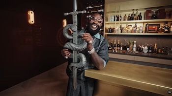 Hombre con barba y gafas de sol sonriendo y sosteniendo una espada con una serpiente, detrás de una barra dorada con estantes llenos de botellas