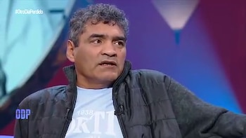 Un hombre de cabello cano y chaleco negro sentado en un estudio de televisión con iluminación azul y violeta