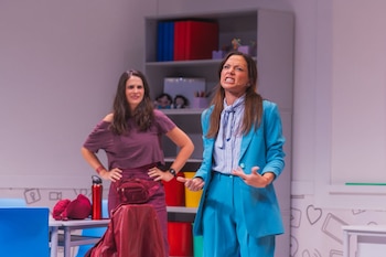 Dos mujeres en un escenario teatral. Una en traje azul turquesa gesticula con enojo, la otra de púrpura, con las manos en la cintura, mira sonriente