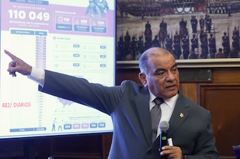 Un hombre de traje gris sostiene un micrófono mientras señala con el dedo hacia una pantalla interactiva con cifras e infografías