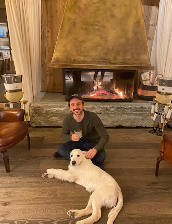 Un hombre sonriente arrodillado en un suelo de madera acaricia a un cachorro de pastor alemán claro que yace frente a él, con una chimenea encendida de fondo