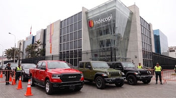 Una camioneta RAM roja y dos vehículos Jeep están estacionados frente a un edificio moderno de vidrio con el logotipo de Indecopi