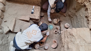 Dos arqueólogos excavan una fosa rodeada de adobes que contiene múltiples vasijas de cerámica y una pieza metálica verdosa