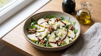 Plato hondo con lonchas de pera, rúcula, nueces, queso desmenuzado, aderezo oscuro y aceite, junto a dos aceiteras sobre una mesa de madera.