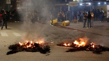 Vista nocturna de una calle de asfalto con dos hogueras ardiendo en primer plano, emanando humo; al fondo, un grupo de personas y barriles amarillos