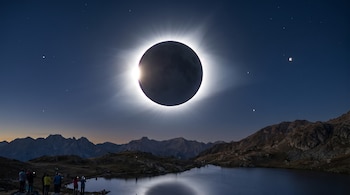 Eclipse solar total en un cielo azul oscuro, con la corona del sol resplandeciendo. Un lago refleja la luz, y montañas rodean a personas observando.