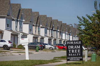 Fila de casas adosadas con tejados a dos aguas, revestimiento claro y porches frontales. Varios automóviles estacionados. Un cartel de “Se Vende” y un buzón blanco