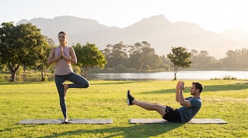 Una mujer en postura de árbol y un hombre en flexión abdominal sobre esterillas en el césped frente a un lago y montañas.