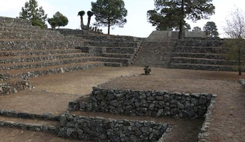 Ruinas de piedra con múltiples terrazas, una escalinata central, paredes de roca, suelo árido y árboles al fondo bajo un cielo nublado