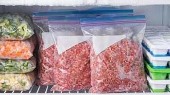 Vista detallada de un congelador con múltiples bolsas de carne molida y vegetales mixtos congelados, apiladas ordenadamente en estantes y recipientes.
