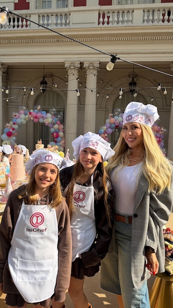 Una mujer y dos niñas con gorros y delantales blancos de cocinero posan frente a una mesa dulce y guirnaldas de globos